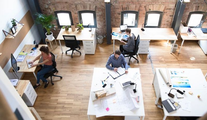 Wide overhead shot of creative office layout with people working at desks adorned with computers, lamps and brochures.